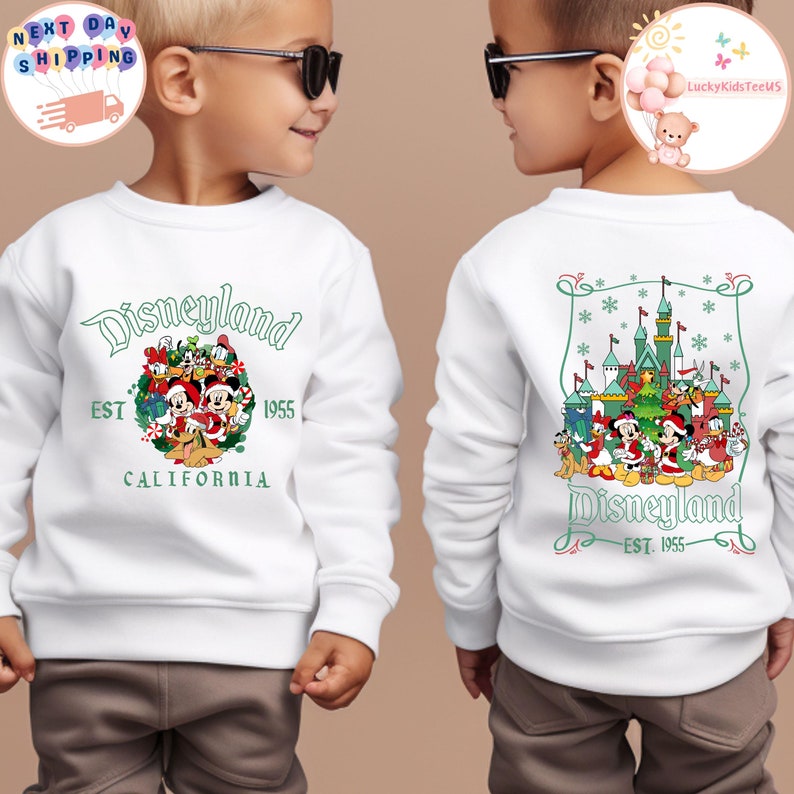 May include: White sweatshirt with a green and red Christmas-themed Disneyland graphic featuring Mickey Mouse and other Disney characters. The text "Disneyland EST 1955 CALIFORNIA" is printed on the front of the sweatshirt.