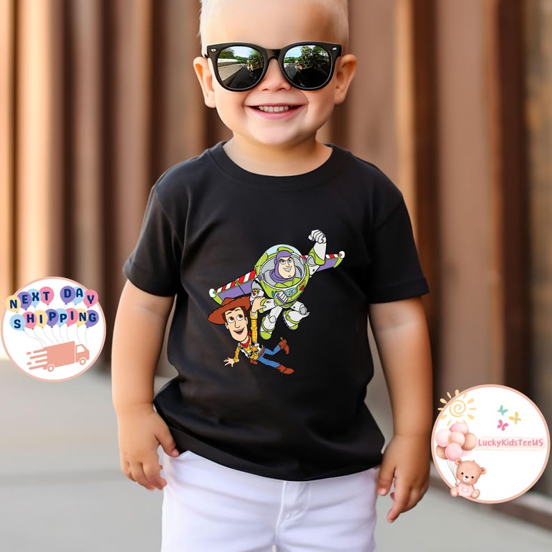 May include: A black t-shirt featuring a colorful graphic of Buzz Lightyear and Woody from Toy Story. The characters are depicted in mid-air. The child is wearing black sunglasses and white shorts.