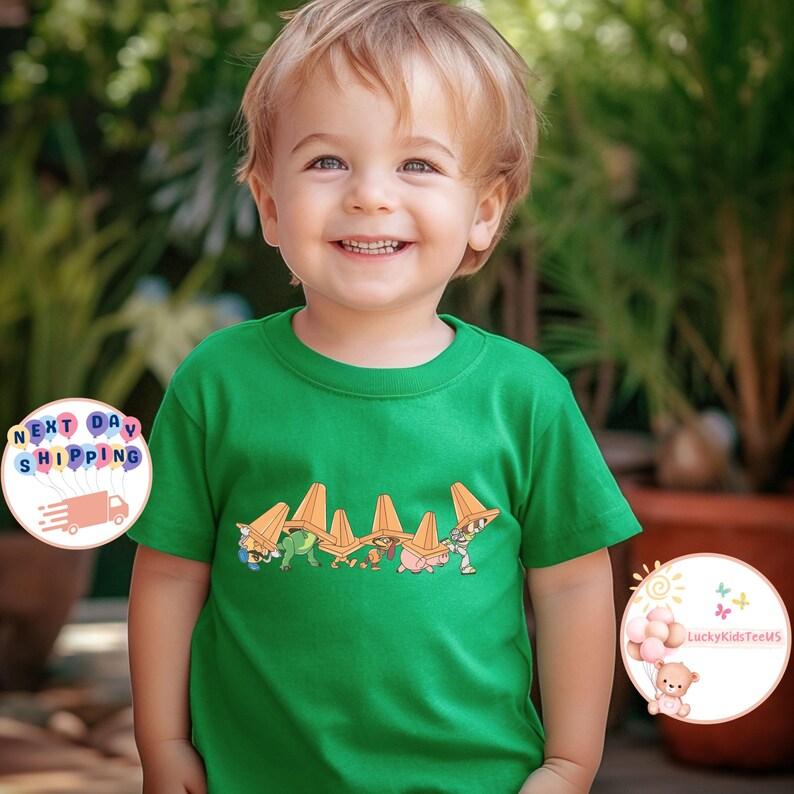 May include: A green crew neck t-shirt featuring a cartoon design of characters wearing orange traffic cones. The design is printed across the front of the shirt. The child is smiling.