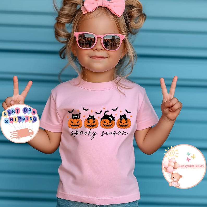 May include: A light pink t-shirt featuring a Halloween design. The shirt displays four orange jack-o'-lanterns with black cat faces and the words "spooky season" in a script font. Black bats are above the pumpkins.