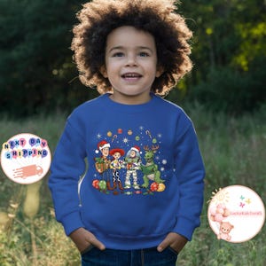 May include: A blue sweatshirt with a Christmas-themed graphic of Toy Story characters, including Woody, Jessie, Buzz Lightyear, and Rex, all wearing Santa hats. The design is surrounded by festive ornaments. The sweatshirt is worn by a young child.