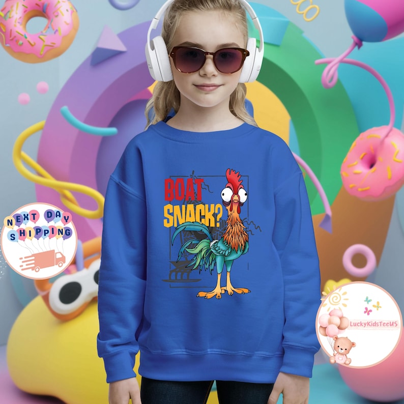 Peut inclure: Un sweat-shirt bleu avec un dessin de coq de dessin anim&eacute; et le texte "BOAT SNACK?" en rouge et jaune. Le coq a une cr&ecirc;te rouge, des plumes bleues et vertes et des pattes jaunes. Le fond comprend des donuts et des formes color&eacute;s.