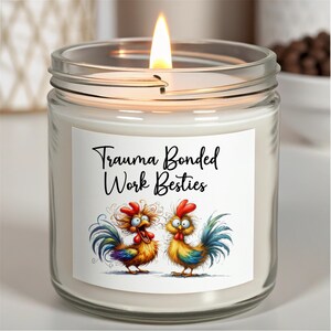 May include: A lit, clear glass jar candle with a white label featuring the text "Trauma Bonded Work Besties" and two cartoon roosters. The roosters are colorful with red combs and blue, yellow, and green tail feathers.