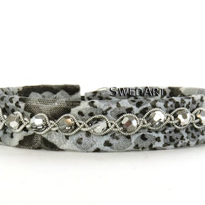 Könnte beinhalten: Ein Armband aus grauem und schwarzem Stoff mit silbernem Draht und klaren Glasperlen. Das Armband hat ein Blumenmuster und das Wort "SWEDART" ist auf dem Stoff sichtbar.