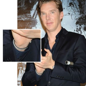 May include: A man wearing a dark blue suit jacket with a white button-down shirt underneath. He is wearing a black bracelet on his left wrist and a black and white bracelet on his right wrist. He is also wearing a black necklace.
