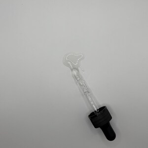 May include: A clear glass dropper bottle with a black cap and a black dropper tip. A small amount of liquid has been dispensed from the bottle onto a white surface.