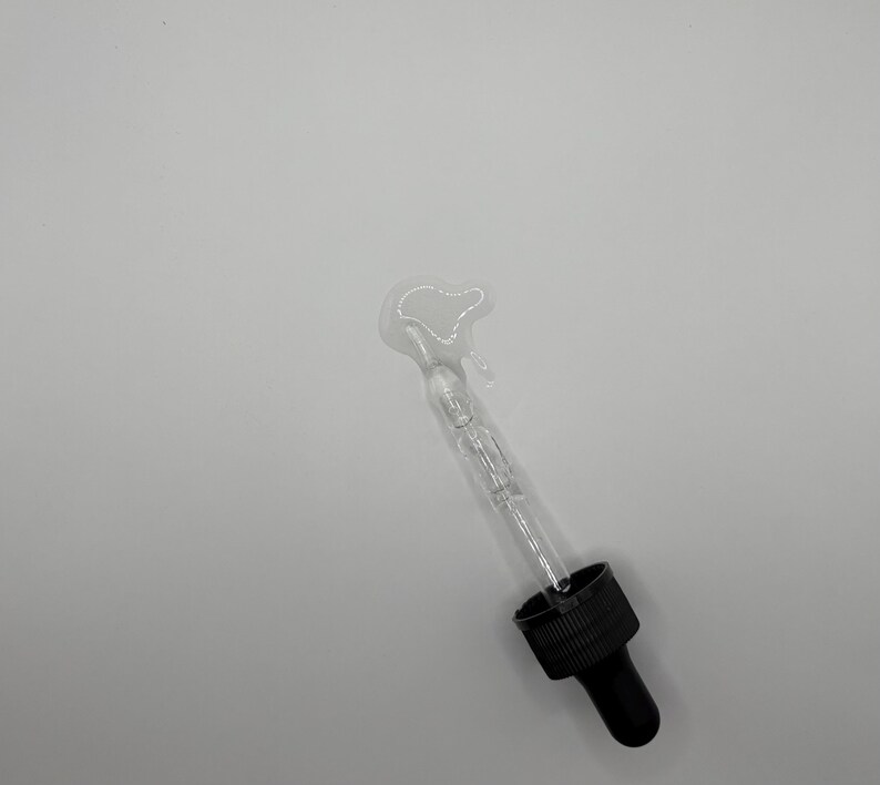 May include: A clear glass dropper bottle with a black cap and a black dropper tip. A small amount of liquid has been dispensed from the bottle onto a white surface.