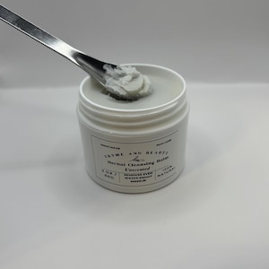 May include: A white jar of Thyme and Beauty Herbal Cleansing Balm, unscented, with a scoop of the balm on a silver spatula. The jar is labeled "HAND MADE SKIN CARE THYME AND BEAUTY Herbal Cleansing Balm Unscented 2 OZ / 60G REMOVES EVEN WATER-PROOF MAKEUP 100% NATURAL"