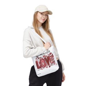 Puede incluir: Bolsa de lona blanca con un diseño gráfico rojo y negro que dice "remarkable love" con corazones y flores. La bolsa está siendo sostenida por una persona que lleva una sudadera blanca y una gorra de béisbol beige.
