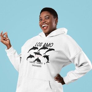 Puede incluir: Sudadera con capucha blanca con un gráfico negro de delfines y el texto "LOS AMO" y "SABIOS SANDS Y SAGRADOS".