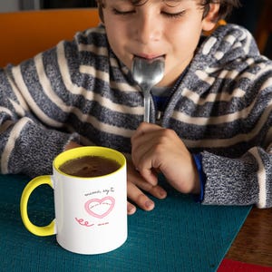 Puede incluir: Una taza de cerámica blanca con un borde y un asa amarillos. La taza tiene un corazón rosa con el texto "mommy says to love you" y "ee" escrito en cursiva. La taza está sobre un mantel individual turquesa.