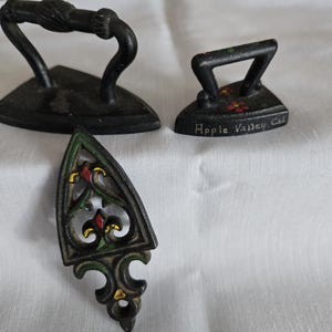 May include: Three antique cast iron miniatures of irons. One has a decorative handle, another has floral accents and the text "Apple Valley, Cal", and the third has a decorative openwork design with red, green, and gold accents.