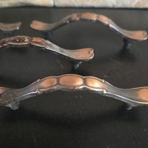 May include: Close-up of several antique bronze cabinet pulls. The pulls have a curved design with decorative detailing and a distressed finish. The hardware is set against a dark surface, highlighting the aged appearance of the metal.