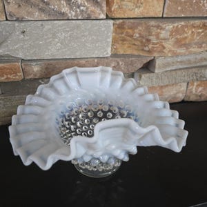 May include: A white, ruffled glass bowl with a hobnail pattern in the center. The bowl has a scalloped edge and sits on a clear, textured base. The bowl is likely vintage or antique.