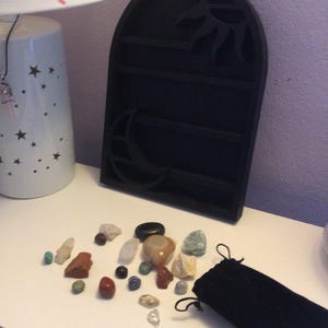 May include: A black wooden display stand with a sun and moon design. The stand has three shelves for displaying small objects. There are various colorful stones and crystals scattered on a white surface next to the stand. A black velvet pouch is also on the surface.