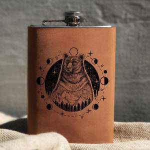 Op de afbeelding: Een bruine leren fles met een zilveren dop. De fles heeft een ontwerp met een beer die in een cirkel van manen en sterren zit.