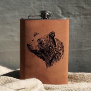 May include: A brown leather flask with a silver top and a black bear design engraved on the front.