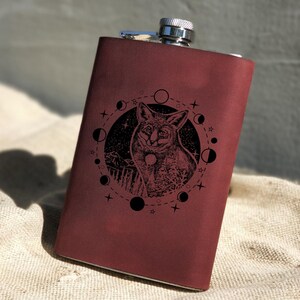 May include: A burgundy leather flask with a silver top and a laser-etched design of a fox in a circle of moons and stars.