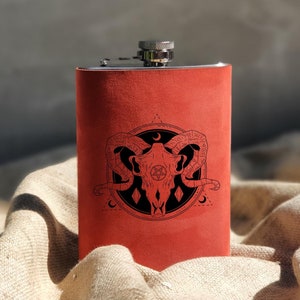 Puede incluir: Una petaca de cuero rojo con tapa plateada y un diseño en blanco y negro de una calavera con cuernos y un pentagrama.