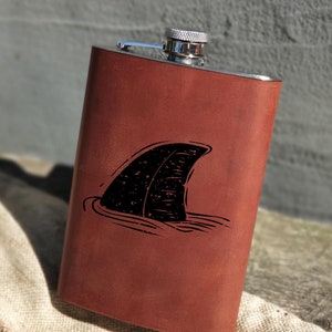 Op de afbeelding: Een bruine leren, roestvrijstalen drinkfles met een gegraveerd zwart haaienvin-ontwerp aan de voorkant.
