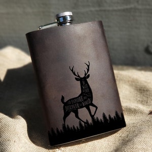 Op de afbeelding: Een bruine leren fles met een zilveren dop en een zwart silhouet van een hert met de tekst "Be wild, explore, discover the world" erop gegraveerd.