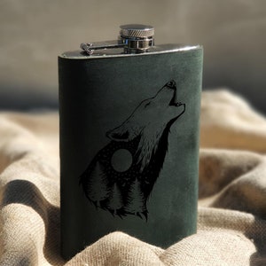 Puede incluir: Una petaca verde oscuro con tapa y bisagra plateadas. Presenta una ilustración negra de un lobo aullando con un diseño de bosque y luna. La petaca es rectangular y parece estar hecha de cuero o un material similar.