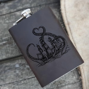 Puede incluir: Una petaca de cuero negro con una tapa plateada y un diseño grabado de un pulpo atacando un barco.