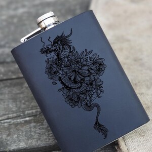 Puede incluir: Una petaca de metal negro con una tapa plateada y un diseño de un dragón rodeado de flores grabado con láser.