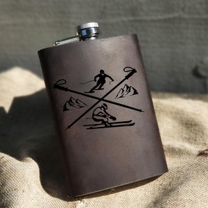 May include: A brown leather flask with a silver cap and a laser engraved design of a skier with ski poles and mountains.