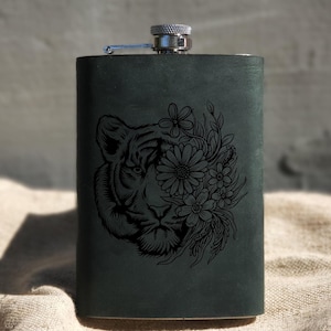 Puede incluir: Una petaca de cuero negro con tapa plateada y un diseño grabado con láser de la cara de un tigre con flores.
