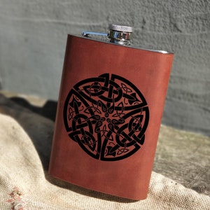 Puede incluir: Una petaca de acero inoxidable envuelta en cuero marrón con una tapa plateada. La petaca presenta un diseño de nudo celta negro.