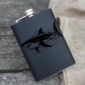 May include: A black metal flask with a shark graphic. The shark is black and white and is swimming in waves. The flask has a silver cap.