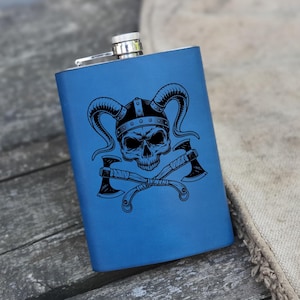 Puede incluir: Una petaca de metal azul con un diseño de calavera negra y hachas cruzadas. La calavera lleva un casco con cuernos.