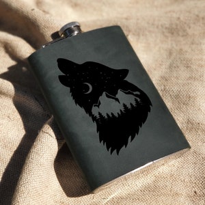 Op de afbeelding: Een zwarte metalen fles met een ontwerp van een wolf die huilt naar de maan. De silhouet van de wolf is gevuld met een bergketen en bomen.