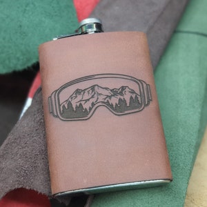 Könnte beinhalten: Eine braune Lederflasche mit silbernem Verschluss. Die Flasche zeigt ein Berg- und Baumdesign in einer Skibrillen-Umrandung. Die rechteckige Flasche ist aus Leder. Der Hintergrund zeigt verschiedene farbige Lederstücke.