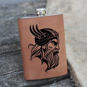 Puede incluir: Una petaca de cuero marrón con tapa plateada y una ilustración en blanco y negro de un guerrero vikingo con casco con cuernos.