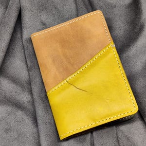 May include: A bi-color leather wallet with a tan suede top and a bright yellow bottom. The two sections are stitched together with white thread. The wallet is resting on a gray, textured surface.