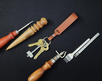 May include: A set of leatherworking tools including a leather punch, an awl, a stitching tool, and a leather strap with a key ring. The tools are laid out on a black surface.