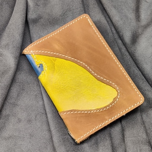 Könnte beinhalten: Ein brauner Leder-Reisepass-Etui mit einem gelben und blauen Ledereinsatz. Das Etui hat einen genähten Rand und einen Klappverschluss.