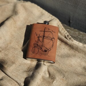 Pode incluir: Uma garrafa de couro marrom com uma tampa de prata e uma gravação detalhada de um navio à vela.