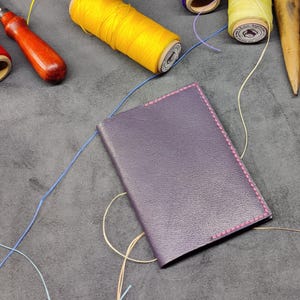 May include: A purple leather notebook with pink stitching, surrounded by spools of thread in various colors, including red, yellow, blue, and white.
