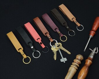 May include: Assortment of eight leather keychains in various colors, including brown, pink, black, and yellow. Each keychain has a metal ring and a leather strap. The keychains are arranged on a black surface.