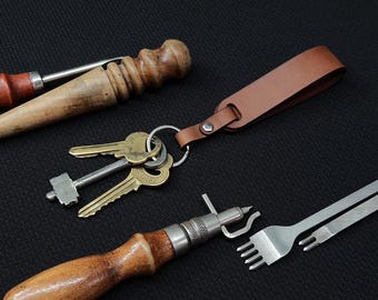 May include: Three leather working tools, including a stitching awl, a stitching pony, and a leather punch. The tools are on a black surface. There is also a set of keys with a brown leather key chain.
