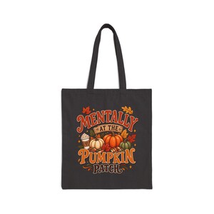 May include: A black tote bag featuring a graphic print with the text "MENTALLY AT THE PUMPKIN PATCH" in orange and white. The design includes illustrations of orange and green pumpkins, a coffee cup, and colorful autumn leaves on a dark background.