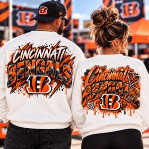 May include: Two white crewneck sweatshirts featuring Cincinnati Bengals graphics. The left sweatshirt displays bold orange and black text with a tiger stripe pattern. The right sweatshirt features orange and black text with heart accents.
