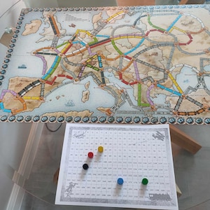 Op de afbeelding: Een spelbord voor het bordspel Ticket to Ride Europe. Het bord toont een kaart van Europa met treinroutes die steden verbinden. Er zijn vier gekleurde speelstukken op een geruit scoreblad.