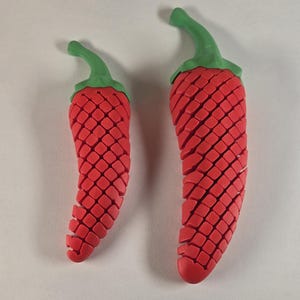 May include: Two red chili pepper shaped ornaments with green tops. The chili peppers are made of a textured material that resembles scales.