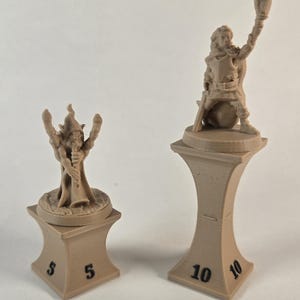 May include: Two miniature figurines on bases, one is a brown elf-like figure with raised arms and the other is a brown human-like figure holding a torch. The bases are brown and have numbers printed on them, 5 and 10.