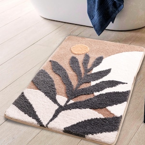 Può includere: Un tappetino da bagno beige, bianco e grigio con un design a foglia stilizzato. Il tappetino ha una superficie testurizzata e un sottile design a sole nell'angolo in alto a destra.