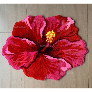 Puede incluir: Una alfombra hecha a mano con forma de flor de hibisco. La alfombra presenta un degradado de colores, con pétalos rojo oscuro que pasan a bordes rosas. El centro tiene estambres amarillos y el texto "Svethomeuk" es visible.